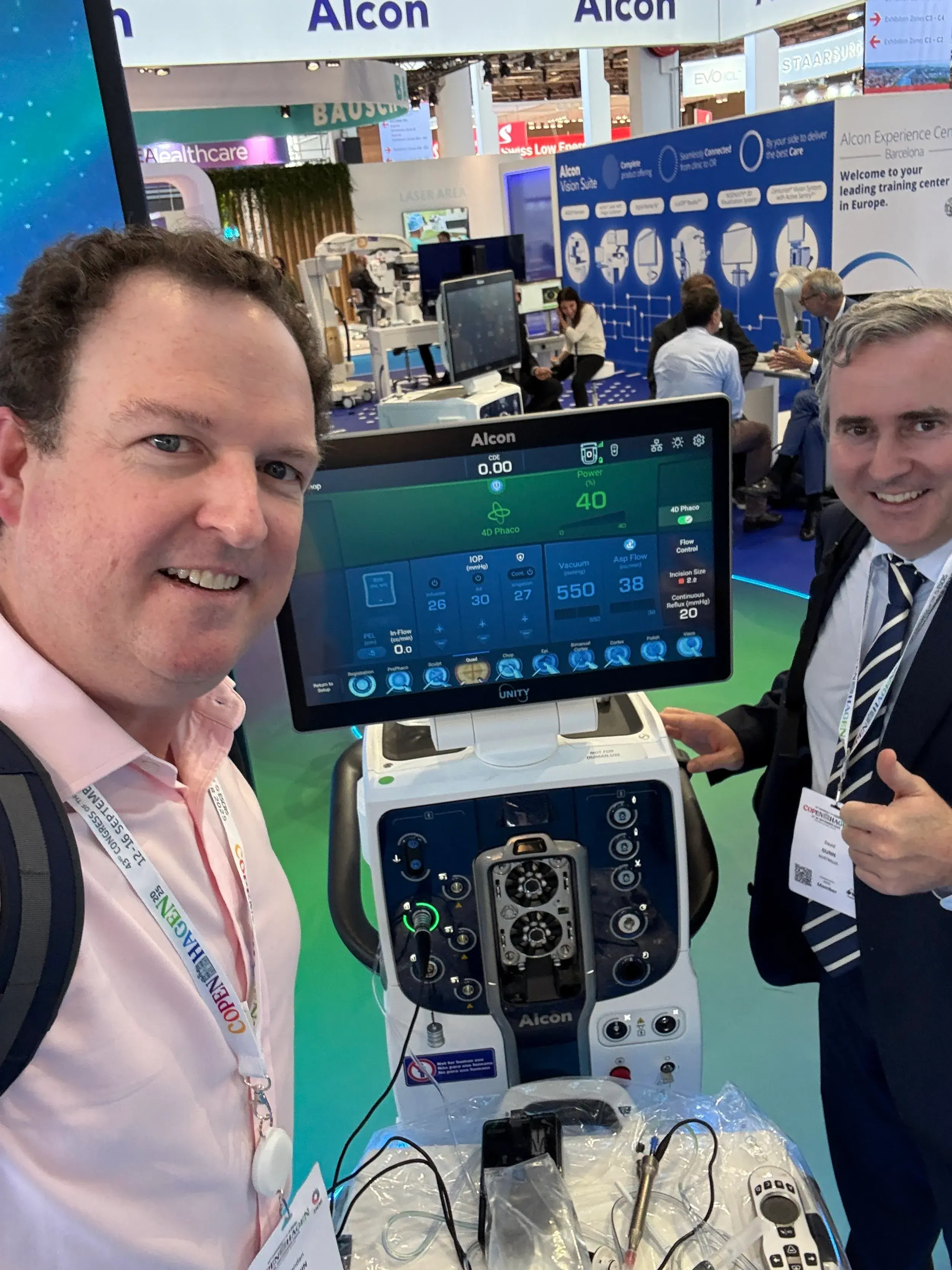 Two men pose in front of Alcon medical equipment at an exhibition.