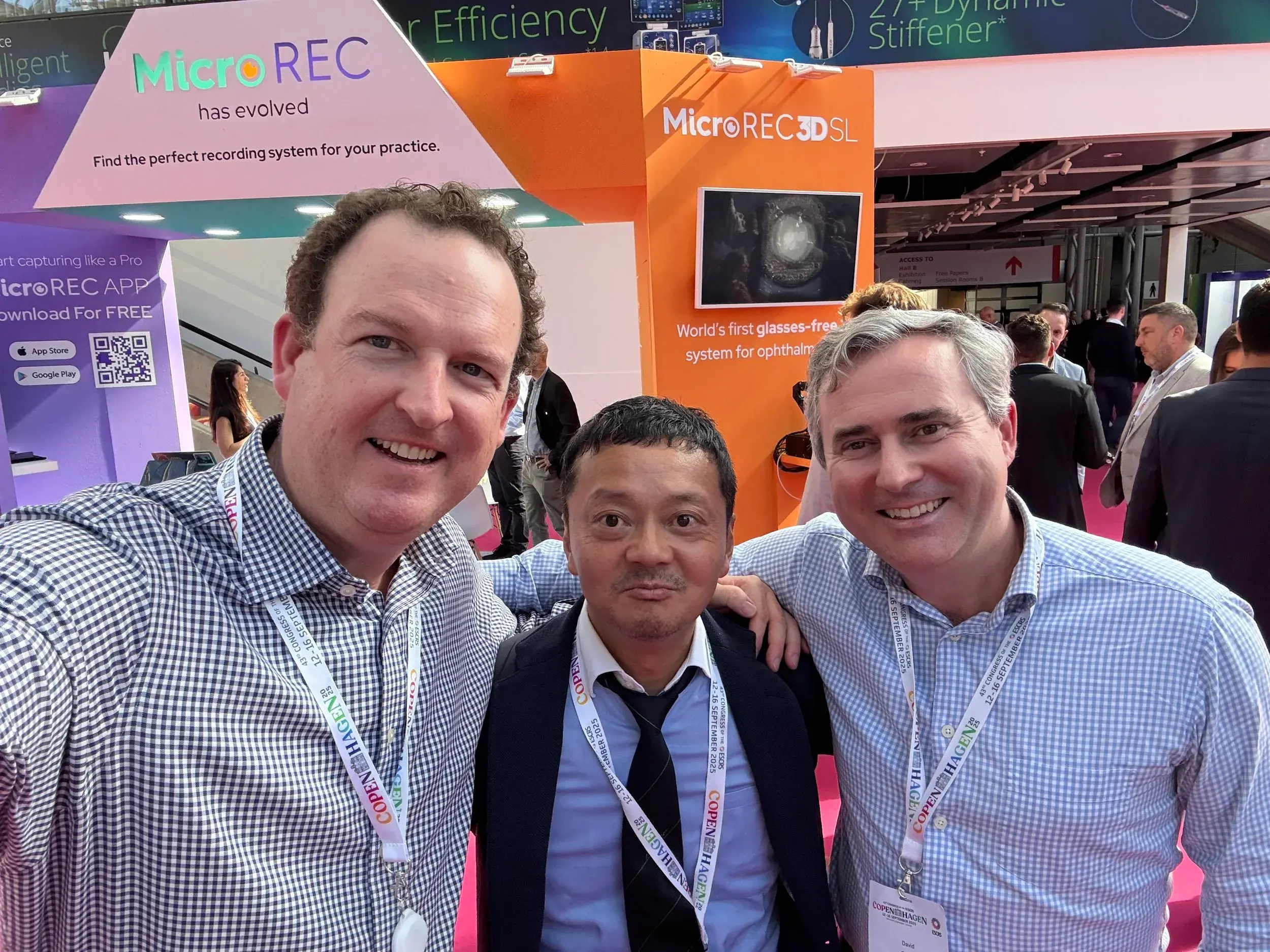 Three men posing for a selfie at the ESCRS conference, smiling with booths in background.
