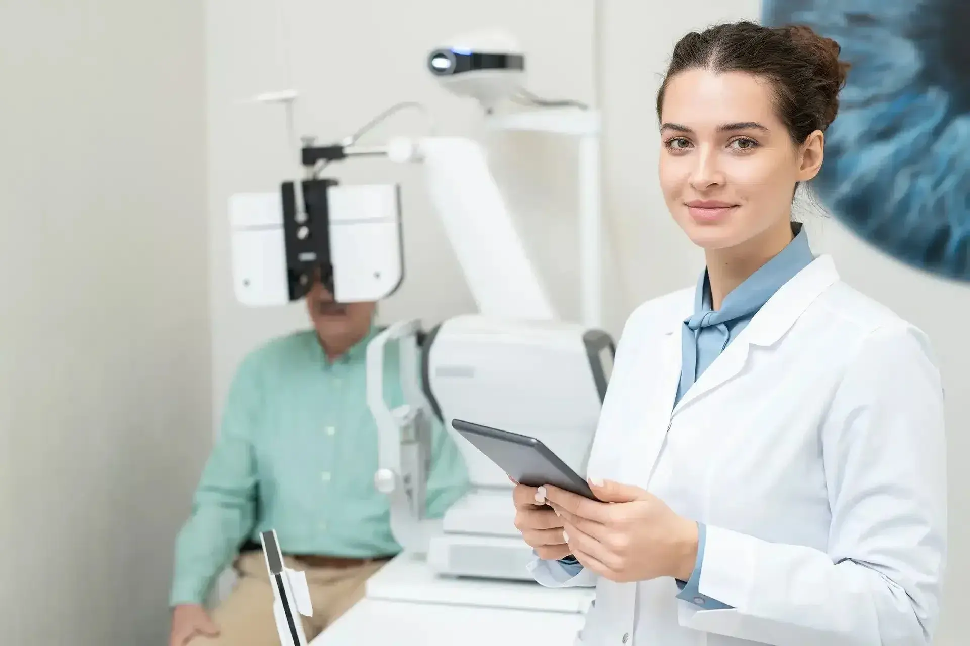 Optometrist assistant holding a tablet, with a patient undergoing an eye exam.