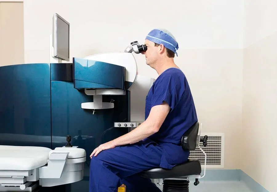 Dr Cronin a corneal surgeon in blue scrubs and a surgical cap sits on a stool, looking into a medical laser machine, likely for an eye procedure, in a clinical setting in Brisbane.