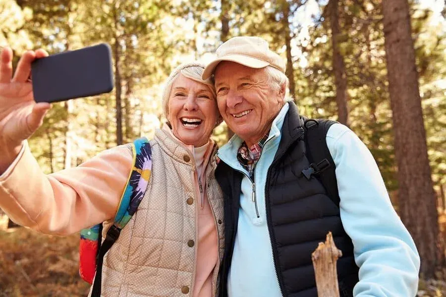 An older couple, smiling and wearing outdoor jackets, takes a selfie together in a sunlit forest. They appear happy after a consultation with Dr Brendan Cronin, surrounded by trees and nature.